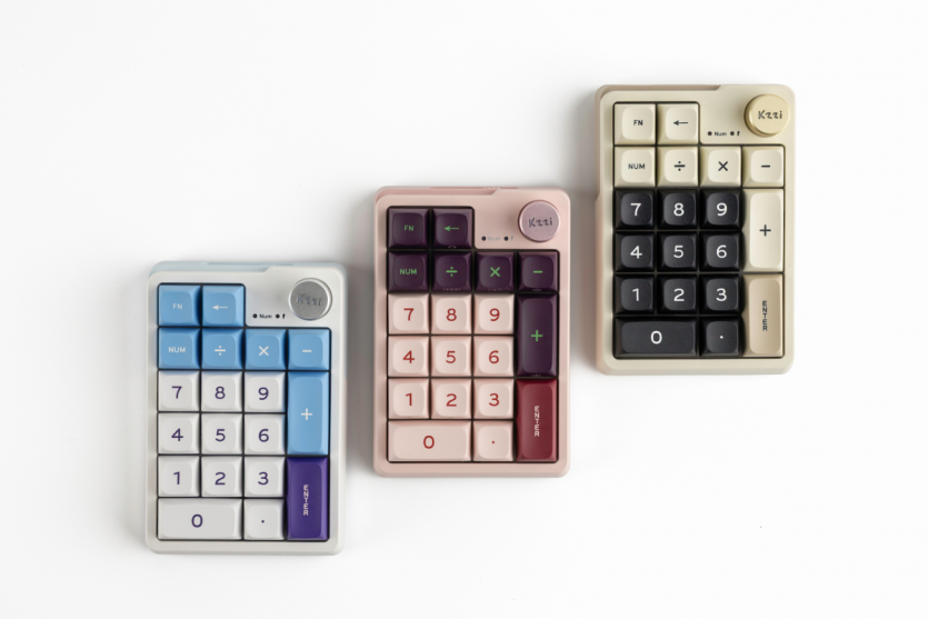Три кольори механічного нампада Kzzi K20: блакитний Blue Sea, рожевий Nezuko Purple та бежевий Starry Gray, що лежать поруч.
